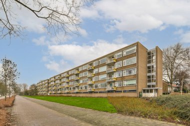 Önünde ağaçlar ve çalılar olan, beyaz bulutlu mavi gökyüzünün altında bir apartman dairesi.