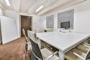 Sandalyeli bir konferans odası ve odanın ortasında büyük beyaz bir masa duvarda bir televizyon var.