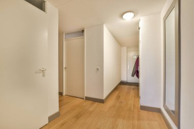 Odanın sağ tarafında beyaz duvarları ve ahşap döşemeleri olan boş bir oda, duvarda bir ayna var.