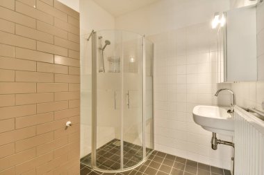 Köşesinde lavabo ve duş kabini olan bir banyo yanında fayanslı bir duvar ve yerde beyaz fayanslar var.