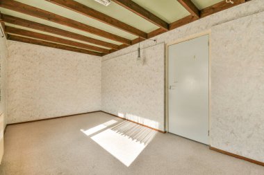 Beyaz duvarları ve tavanında tahta kirişleri olan boş bir oda. Kapıdan içeri ışık giriyor.