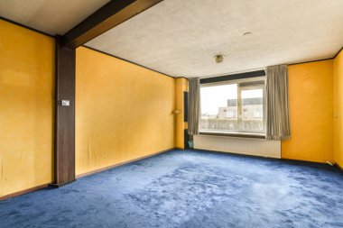 Odanın ortasında sarı duvarları ve mavi halısı olan boş bir oda var.