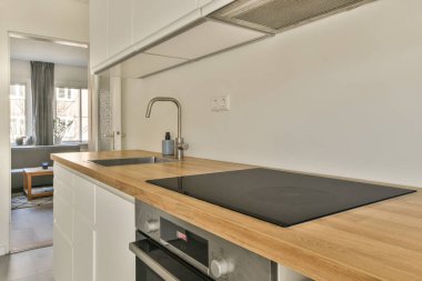 Ahşap tezgahlı ve arkasında beyaz dolaplar olan bir mutfak. Açık bir kapı oturma odasına açılıyor.