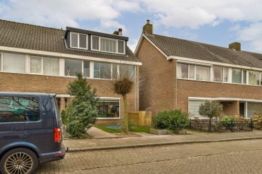 Ağaçları ve çalıları olan kaldırımlı bir caddede tuğlalı evlerin önüne park etmiş bir araba.