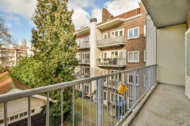 Arka planda ağaçlar ve binalar olan bir balkon. Bir apartmanın penceresinden sokağa bakıyor.