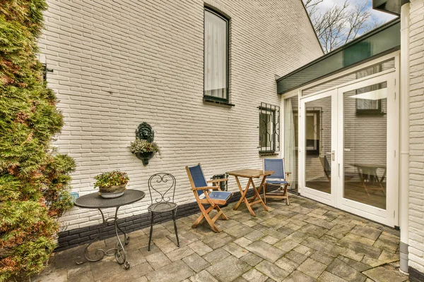 a patio with two chairs and a table in front of a white brick wall that has green plants growing on it