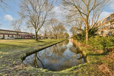 Yerleşim alanının ortasında küçük bir gölet, çıplak ağaçlar ve yerde yaprak ve çimen yok.