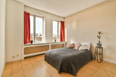 Penceresinde kırmızı perdeler olan bir odada bir yatak açık ve yanında bir bitki var.