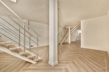 Odanın ortasında ahşap döşemeli ve merdivenli boş bir oda. Sağ tarafta beyaz bir duvar var.