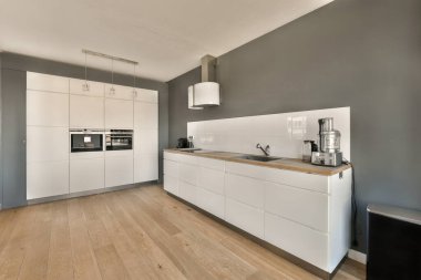 Ahşap döşemeli bir mutfak ve arkasındaki duvarda beyaz dolaplar bir fırın, mikrodalga fırın ve bulaşık makinesi var.