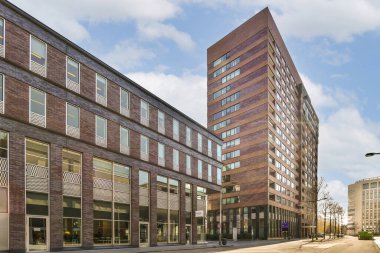 Caddenin kenarındaki tuğladan bir bina. Önünde arabalar, arkasında da insanlar var.