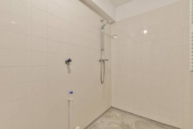 Duvarlarında beyaz fayanslar olan bir banyo ve duvara monte edilmiş duş başlığı köşedeki tuvaletin yanında.