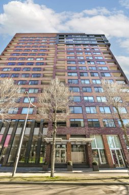 Önünde birçok penceresi olan yüksek bir bina ve arkasında mavi bir gökyüzünün altında yanan ağaçlar.