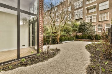 Fotoğrafın ortasında ağaçlar ve bitkiler olan bir dış alan. Diğer tarafında tuğla bir bina var.