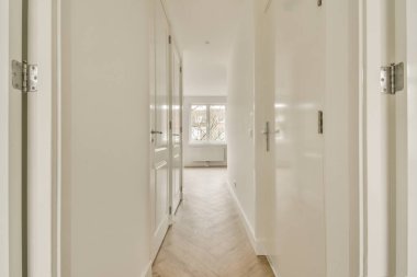 Her iki tarafında beyaz duvarlar ve ahşap döşemeler olan uzun bir koridor, başka bir odaya açılan açık bir kapıya çıkıyor.