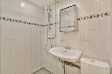 Duvarlarında beyaz fayanslar ve yeşil süslemeler olan küçük bir banyo. Lavabonun üstünde asılı bir ayna var.