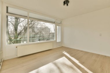 Boş bir oturma odası, ahşap döşemeleri ve geniş pencereleri var. Uzaktaki ağaçlara bakan çıplak dallar.