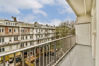 Arka planda bazı binalar ve üzerinde mavi gökyüzü olan bir balkon, bir apartman penceresinden görüldüğü gibi.