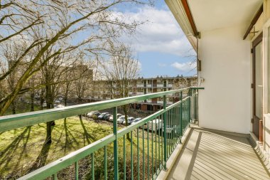 Arka planda yeşil korkuluklu bir balkon ve güneşli bir günde bir apartmanın penceresinden alınan ağaçlar.