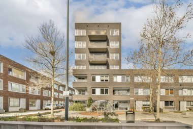 Ön planda ağaçlar ve caddelerde yan yana park edilmiş arabalar olan bir apartman kompleksi.