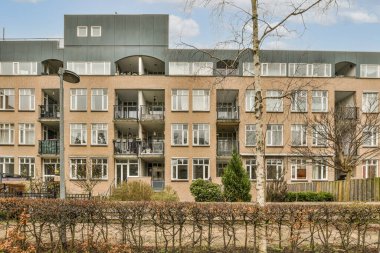 Ön planda ağaçlar ve çalılar olan bir apartman dairesi. Çitin karşısından caddeye götürülmüş.