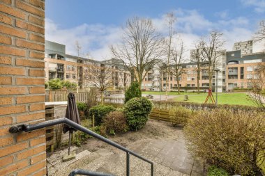 Ön planda ağaçlar ve çalılar olan bir dış alan. Pencereden bazı binalara bakıyor.