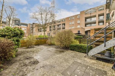 Ön planda ağaçlar ve çalılar olan bir dış alan, apartmana çıkan merdivenlerden görüldüğü gibi.