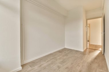 Beyaz duvarları ve sağ tarafında ahşap döşemeleri olan boş bir oda, sol köşede bir kapı var.