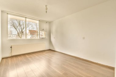 Ahşap döşemesi ve duvarlarında beyaz boya olan boş bir oda. Köşesinde bir pencere var.