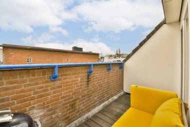 Beyaz tuğlalı bir binanın önündeki duvarda sarı kanepeler ve mavi borular olan bir balkon, parlak mavi bir gökyüzüne karşı.