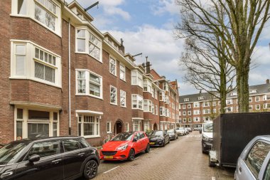 Beyaz pencereli tuğlalı apartmanların önüne park etmiş birkaç araba var.