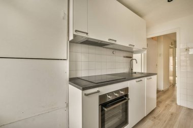 Resmin ortasında beyaz dolapları ve siyah tezgahları olan bir mutfak, başka bir odaya açılan açık bir kapıdır.