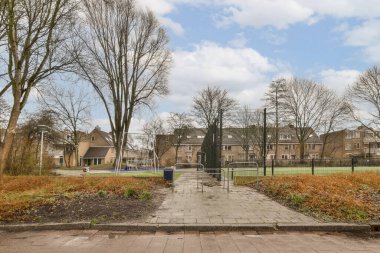 Güneşli bir günde, arka planda ağaçlar ve binalar olan boş bir park, sokağın karşısından görüldüğü gibi.