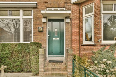 Binanın yan tarafında büyüyen beyaz pencereli ve bitkileri olan kırmızı tuğlalı bir evin önünde yeşil bir kapı.