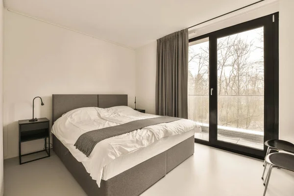 stock image a bedroom with a bed, desk and large window looking out onto the trees in the photo is black and white