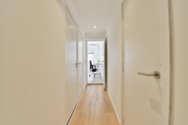 Beyaz duvarları ve yan yana ahşap döşemeleri olan uzun bir koridor. Başka bir odaya açılan bir kapı var.