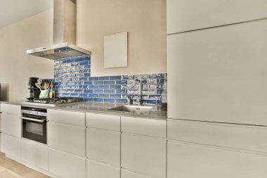 Beyaz dolaplı bir mutfak ve arkasındaki duvarda mavi fayanslar bir fırın, bulaşık makinesi ve lavabo var.