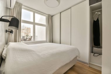 Arka planda yatak odası ve dolapları olan bir yatak odası. Sokağa bakan büyük bir pencere var.