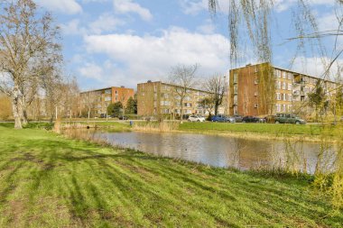Bazı binalar ve sular yeşil çimenler, ağaçlar ve üzerinde mavi gökyüzü olan kentsel bir alanın ortasında.