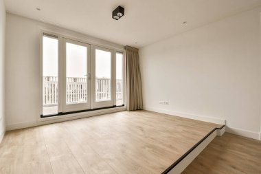 Beyaz duvarları ve ahşap döşemeleri olan boş bir oda. Oturma odasının kapısından balkona bakıyor.