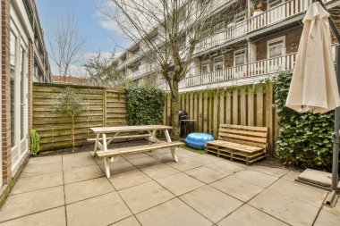 Ağaçlarla çevrili bir apartmanın önündeki verandada bankları, masaları ve şemsiyeleri olan bir arka bahçe.