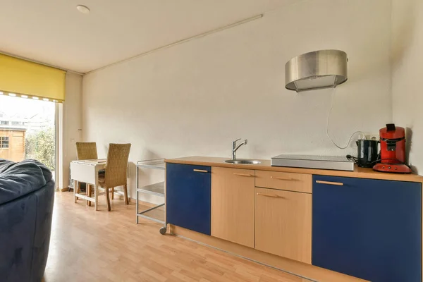 a kitchen and living room in an apartment with wood flooring, blue cabinets, white walls and yellow accent wall