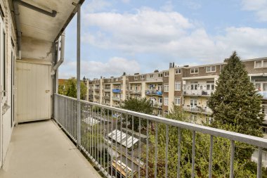 Arka planda ağaçlar ve binalar olan bir balkon. Bir apartmanın içinden alınıp sokağa bakılıyor.