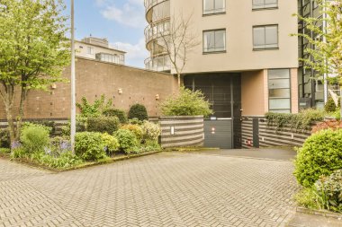 Ağaçları, çalıları ve bitkileri olan bir dış alan tuğlalı bir binanın önünde kırmızı bir yangın musluğu var.