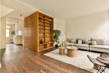 Yerde ahşap döşemeleri ve beyaz halıları olan bir oturma odası, açık bir kitaplık var.