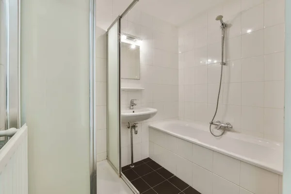 バスタブの中に黒いタイル張りの床と壁にガラスシャワーストールドアの白いバスルームがシンクの隣にあります — ストック写真
