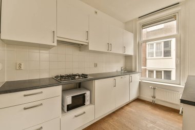 Sobanın önündeki adada beyaz dolapları ve siyah tezgahları olan bir mutfak, yemek pişirmek için kullanılıyor.