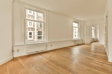 Odanın ortasında ahşap döşemeli ve büyük pencereli boş bir oda beyaz duvarlar, ahşap zeminler.