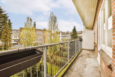 Güneşli bir günde arka planda ağaçlar ve binalar olan bir balkon, bir apartman penceresinden görüldüğü gibi.