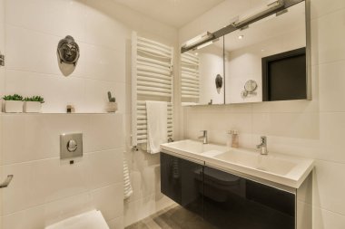 Modern bir banyo, beyaz fayanslar, siyah tezgahlar ve lavabonun üstündeki duvara monte edilmiş bir televizyon.